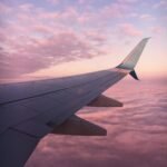 A serene view of an airplane wing flying above clouds during a pink sunset.